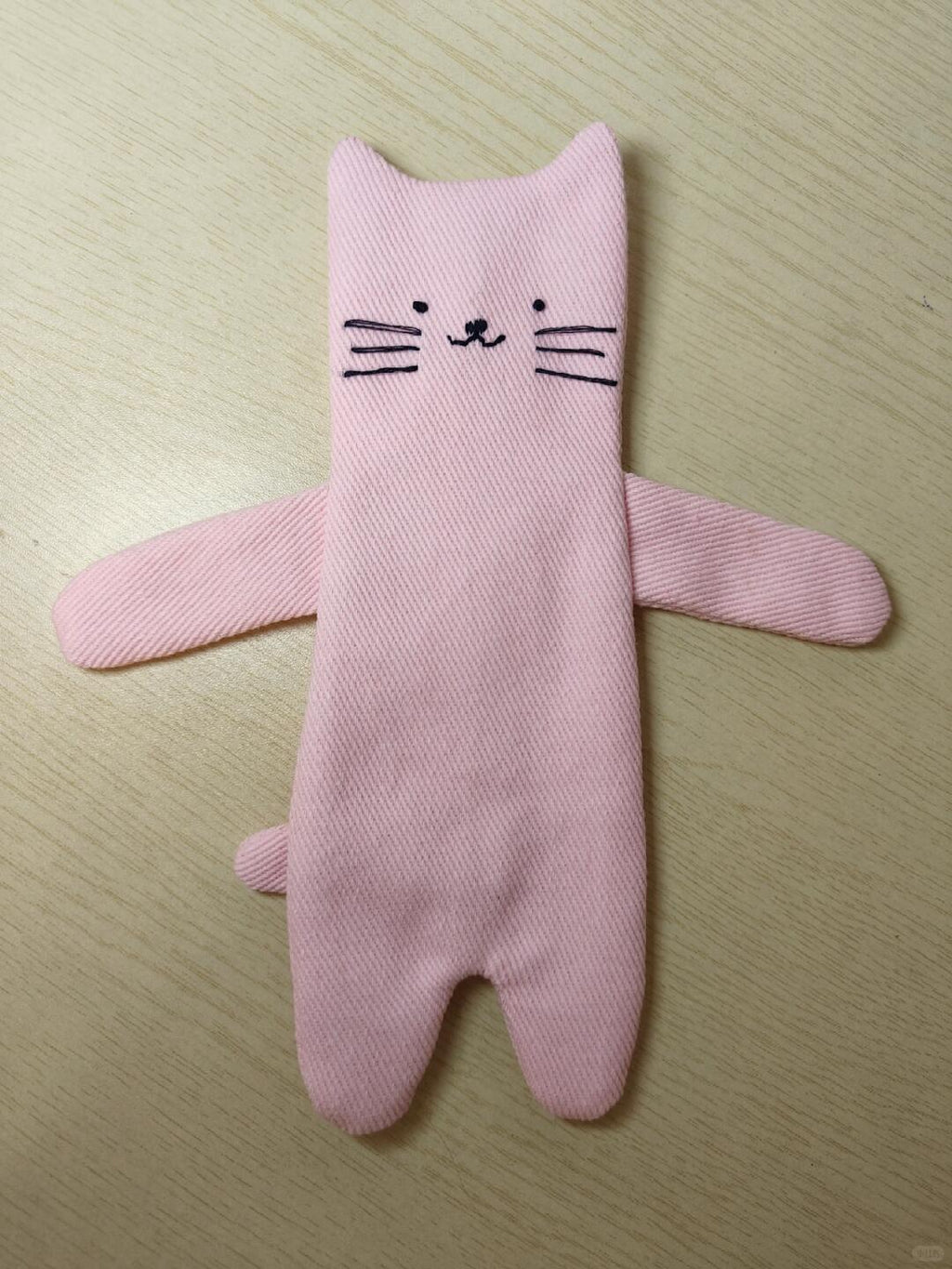 A pink cat-shaped fabric footie sock with embroidered black facial features, displayed flat on a light wood surface.