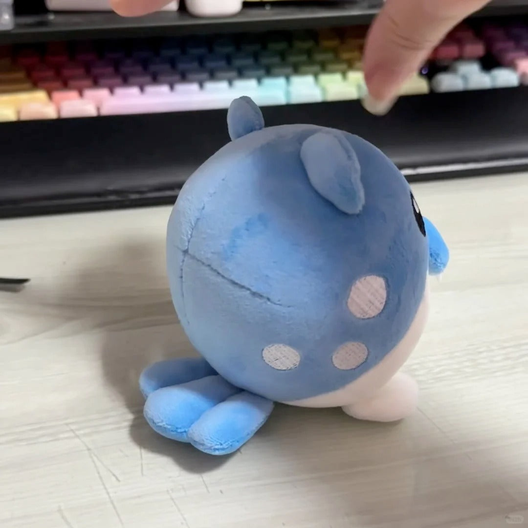 A blue Spheal plush toy with white spots and small ear tufts, photographed on a light tabletop with a keyboard and a finger visible in the blurred background.