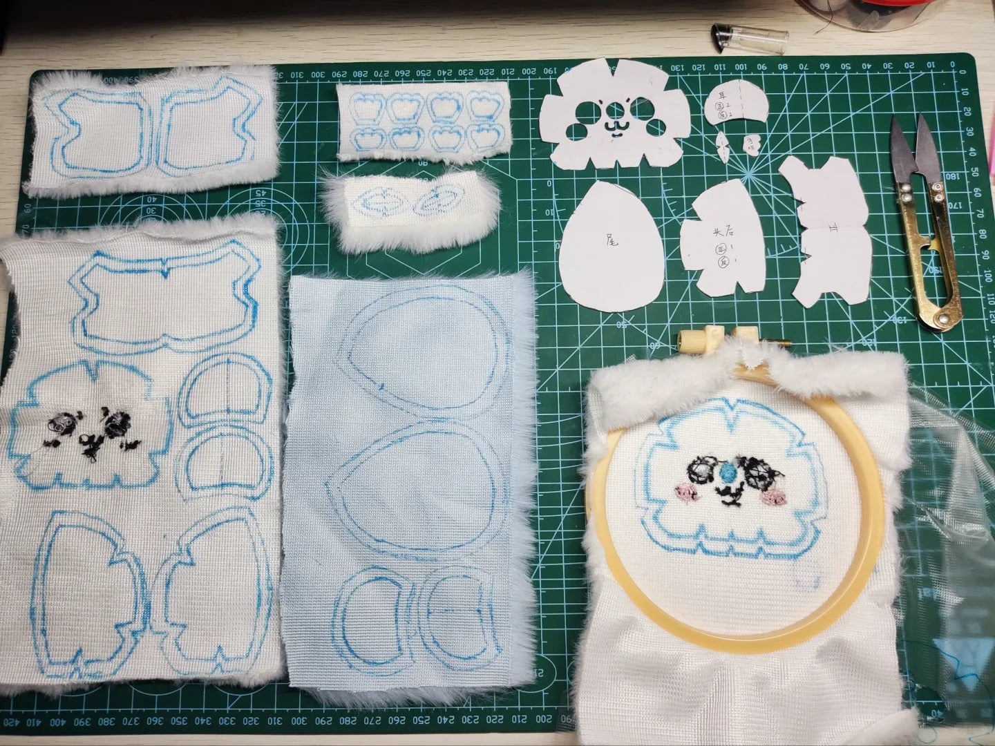Embroidered momonga plush sewing pattern, fabric pieces for a cartoon character, laid out on a green cutting mat alongside embroidery templates, scissors, and a small finished embroidery sample.