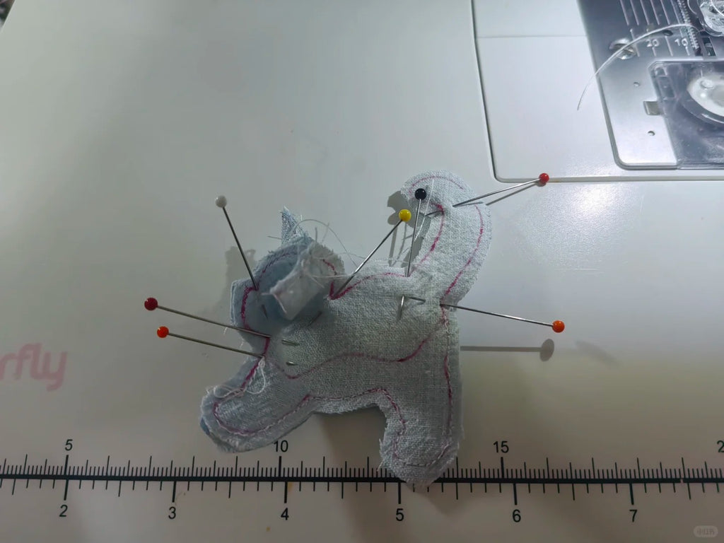 A small handmade fabric pincushion shaped like a cat, pinned with metal pins and red tips, placed on a white sewing machine surface with a ruler visible at the bottom.