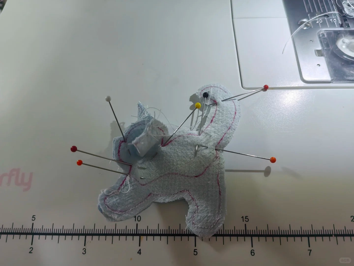 A small handmade fabric pincushion shaped like a cat, pinned with metal pins and red tips, placed on a white sewing machine surface with a ruler visible at the bottom.