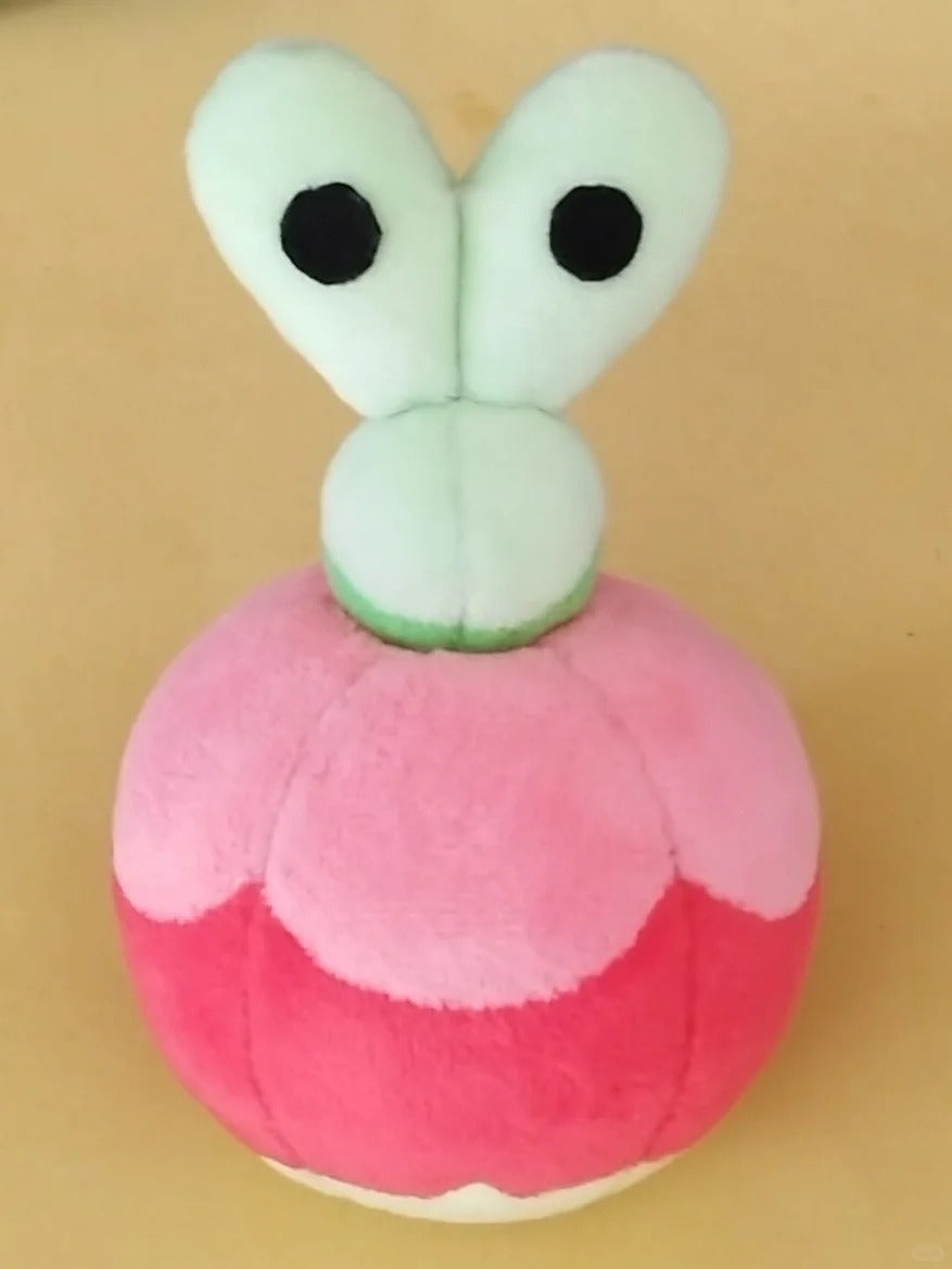 A Applin plush toy shaped like a round pink and coral-pink character head with two large pale mint-green eyes and black dot pupils, photographed against a plain tan background.