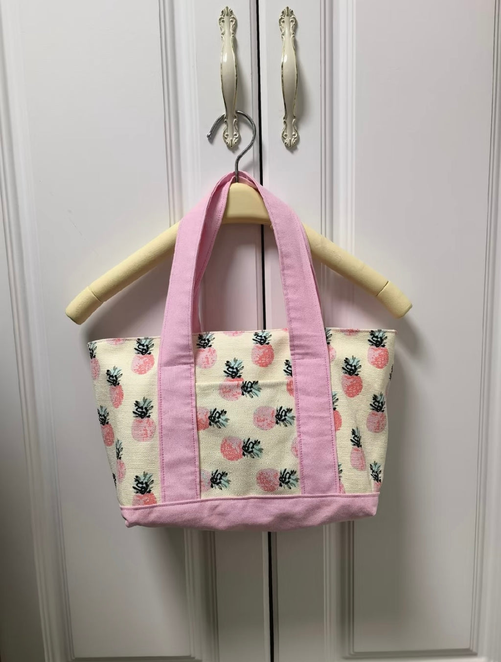 Pink tote bag with pineapple pattern hanging on a wooden hanger against a white wall.