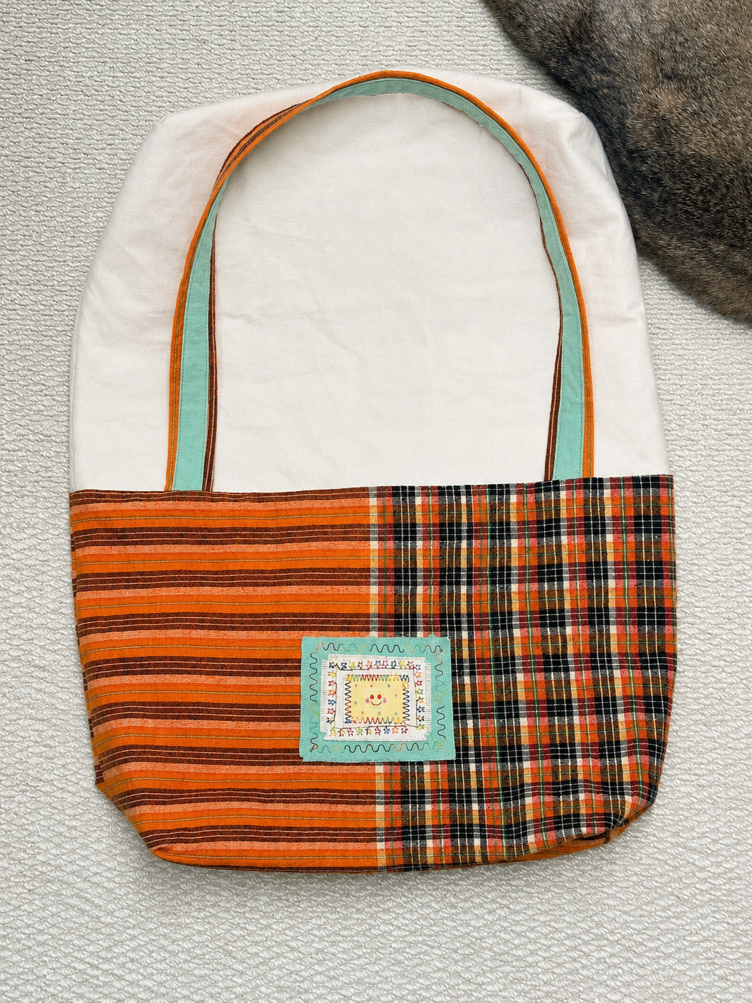 A handmade fabric tote bag with a white canvas upper section, orange striped fabric on the sides, and an orange plaid front panel. It features mint green handles and a decorative square patch on the front.