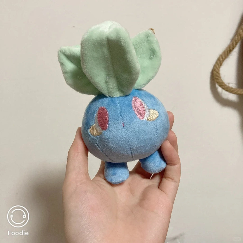 A small blue oddish plush toy with mint green leaf-like structures on top and embroidered pink and cream facial details, being held in a person's hand against a plain indoor wall.