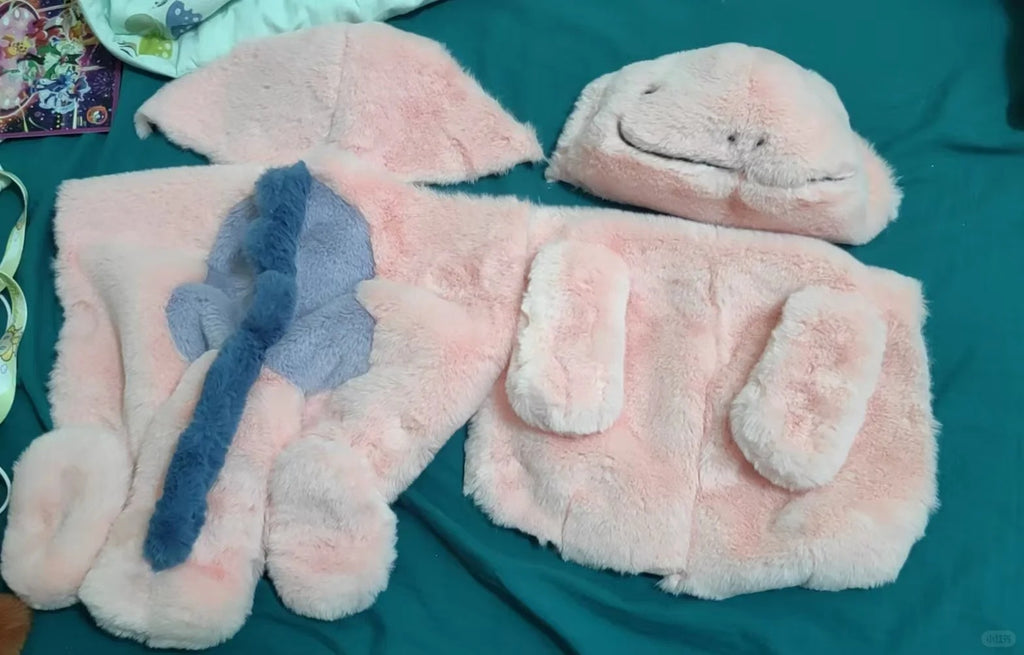 A plush pink fuzzy Quagsire costume or outfit laid out on a teal fabric background, including a hooded headpiece, a face mask, and several matching soft pieces with a blue bow detail.