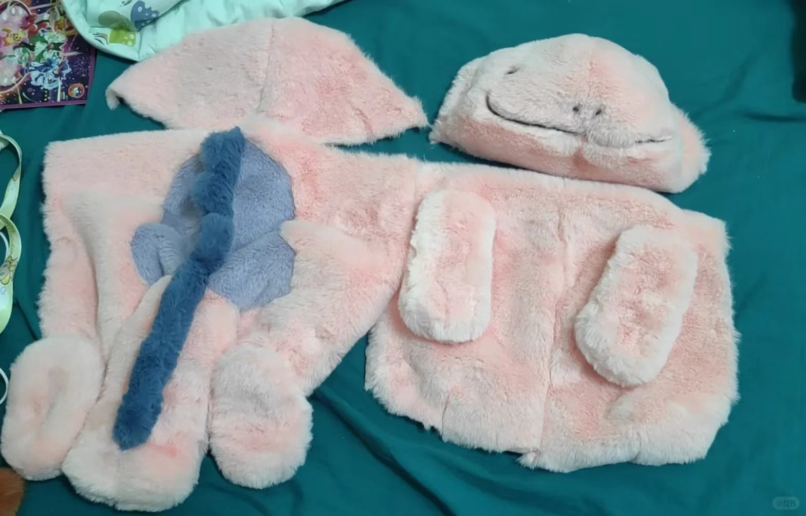A plush pink fuzzy Quagsire costume or outfit laid out on a teal fabric background, including a hooded headpiece, a face mask, and several matching soft pieces with a blue bow detail.