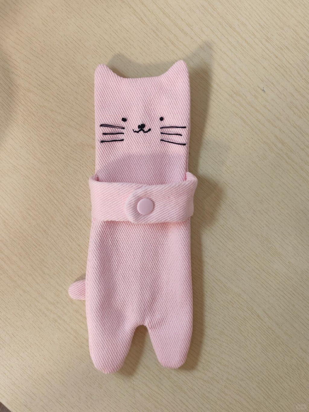 A pink fabric cat-shaped pouch or holder with embroidered cat face details and a snap closure, shown on a light wood surface.