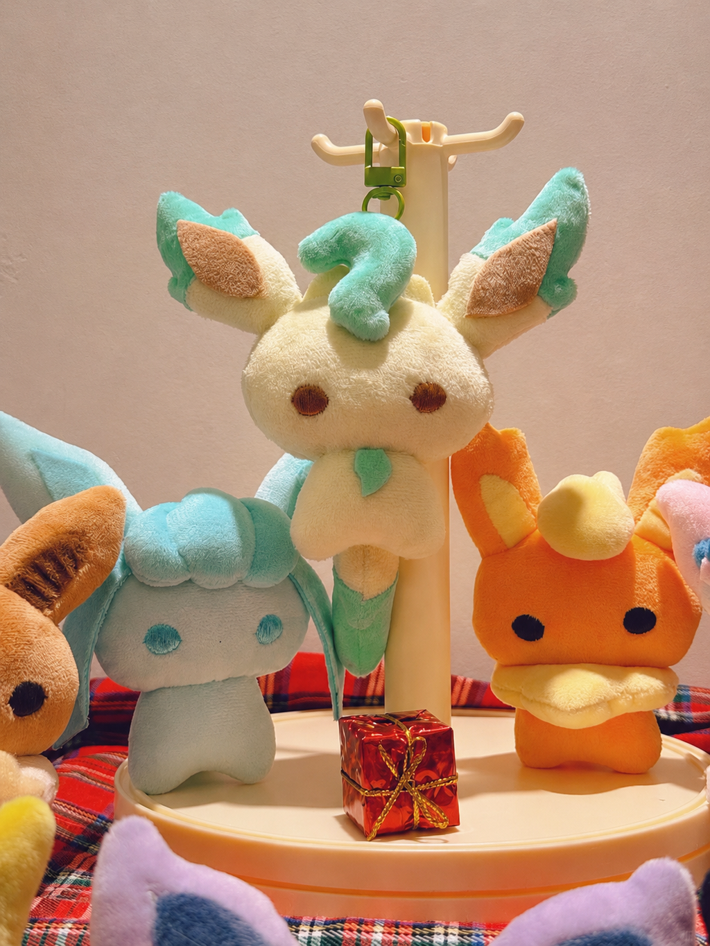 A leafeon, flareon, glaceon, display featuring a central cream-colored Voiding plush with mint green and teal accents, flanked by other colorful plush toys on a beige stand against a plain beige background.