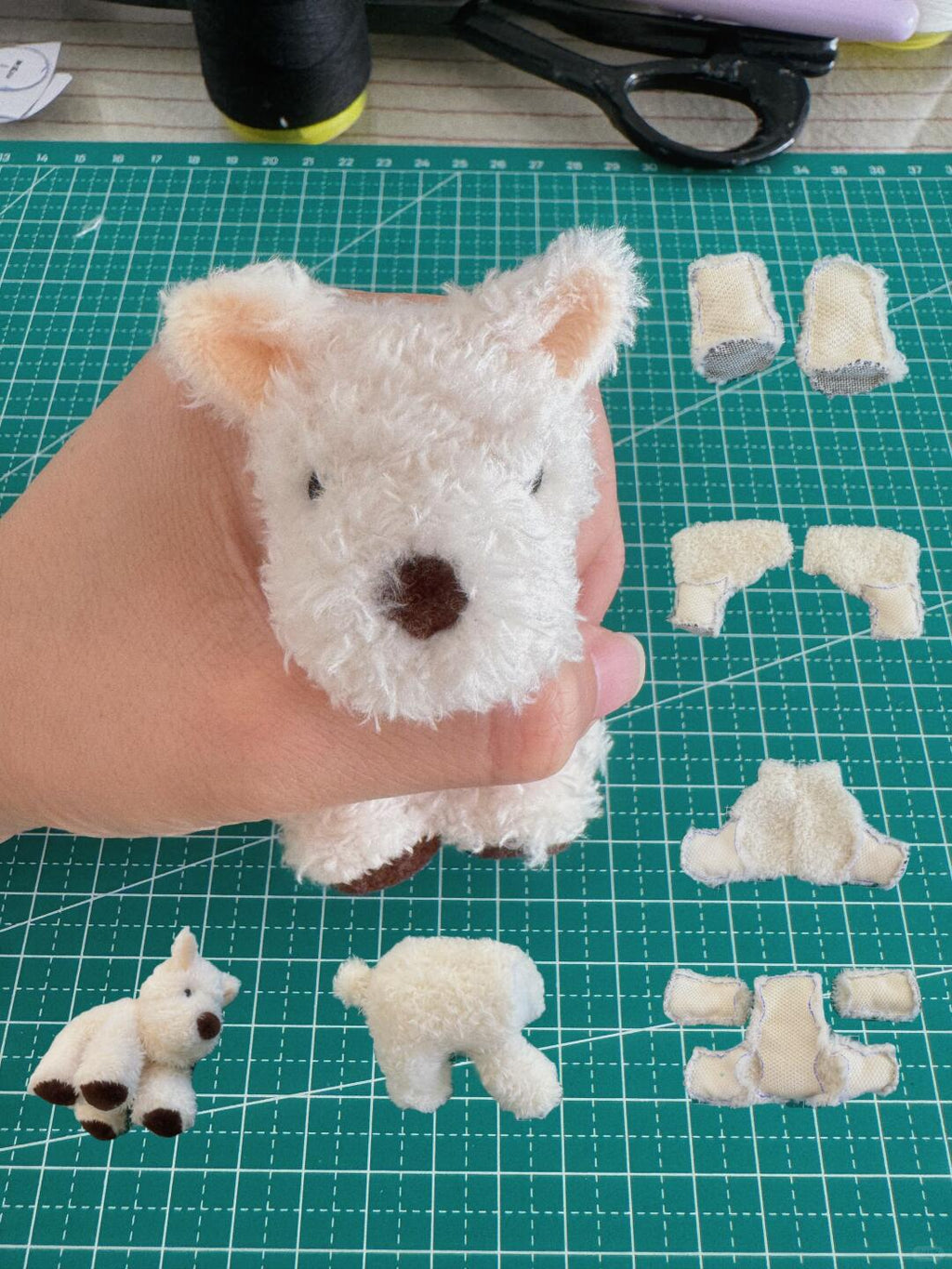 Hand holding a small white teddy bear with cut-out pieces on a green cutting mat.