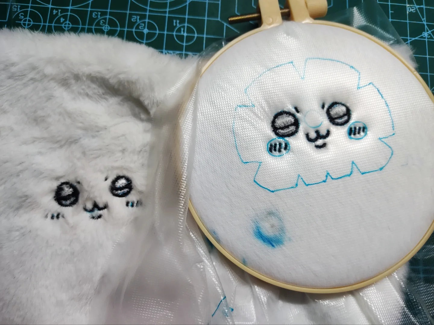 A close-up of a white plush fabric item with embroidered cartoon eyes and mouth, shown next to a wooden embroidery hoop holding white fabric with a blue outline of the same face. A green cutting mat is visible in the background.