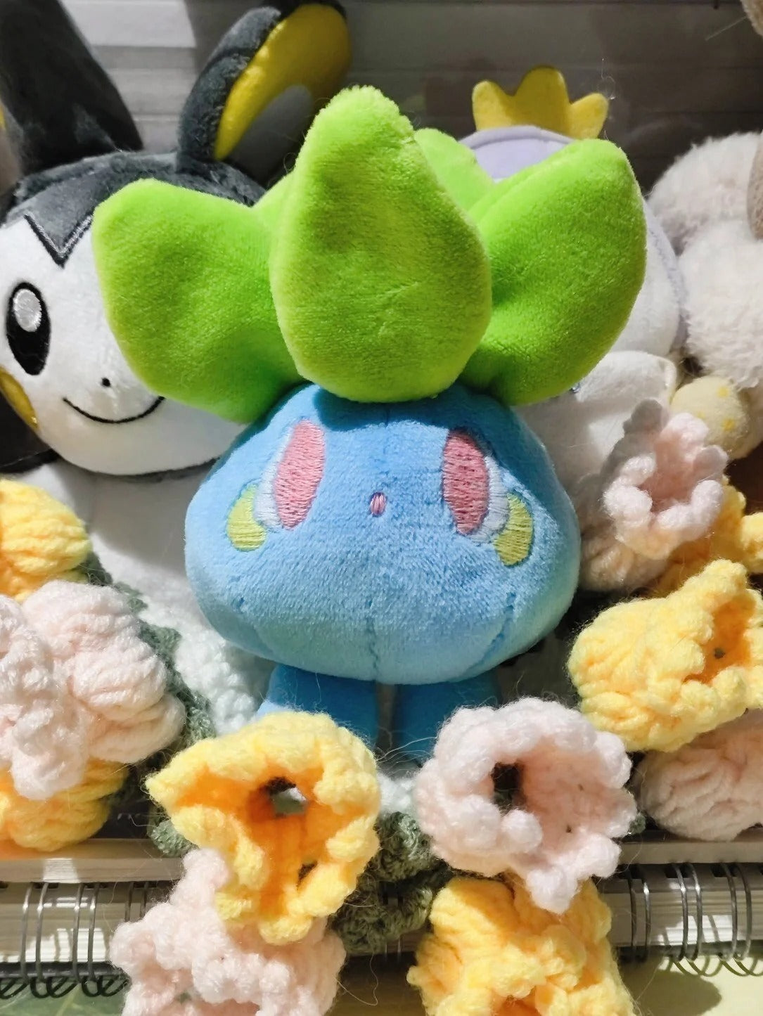 A blue and green oddish pokemon plush toy centered among crocheted flowers, with other plush toys partially visible around it.