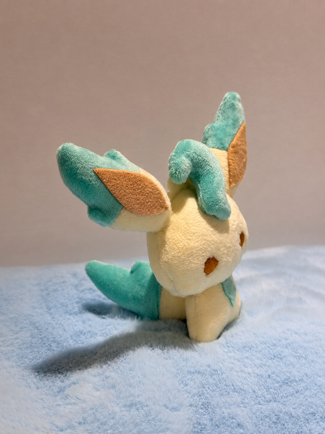 A small leafeon eevee plush toy resembling a cute cream-colored creature with teal and light brown accents, sitting on a soft blue textured surface against a plain beige background.
