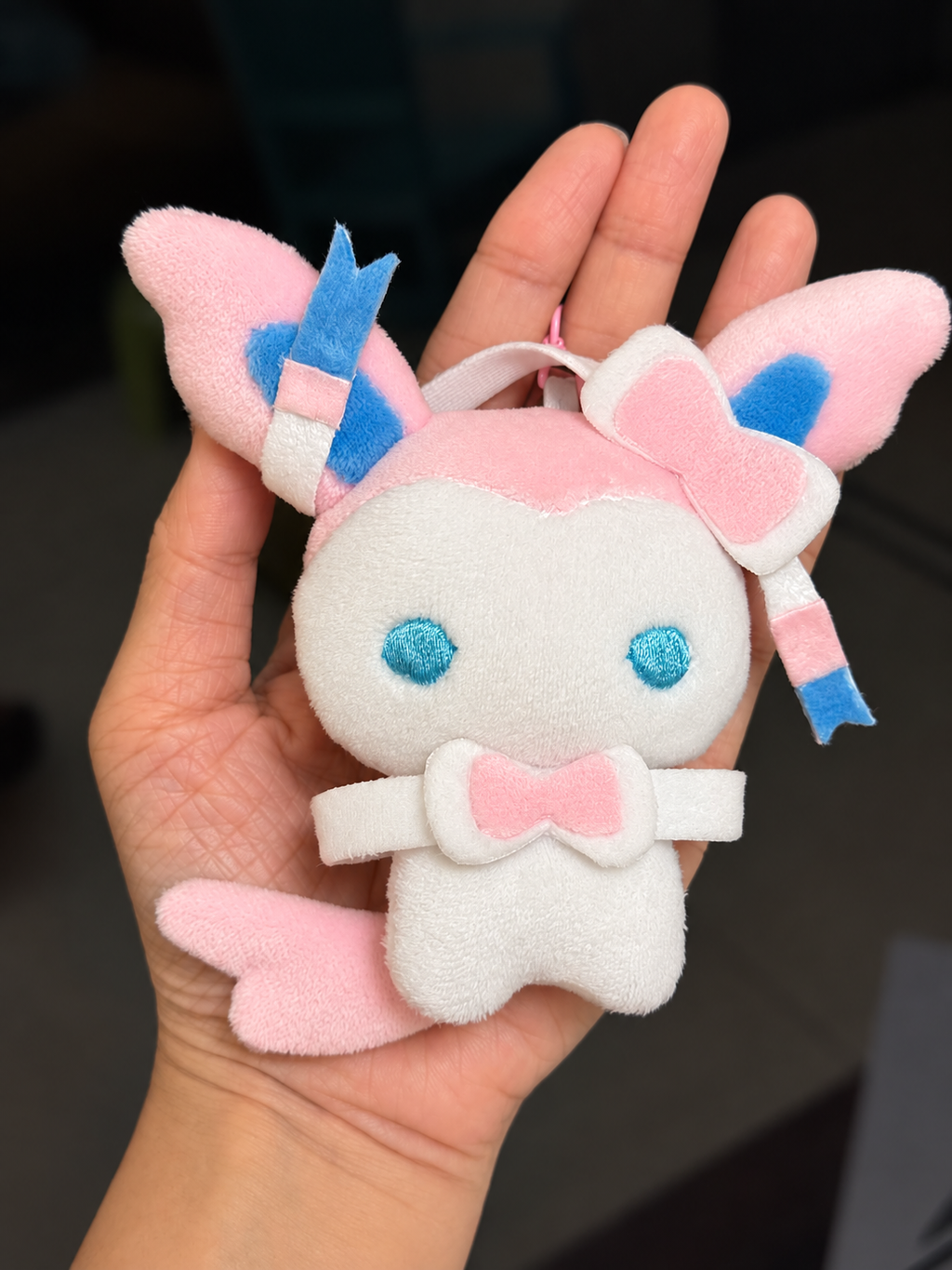 A small plush eevee character keychain or ornament held in a person's hand. The character is white with pink accents, blue embroidered eyes, and pink-and-blue striped ribbon details on its ears and bow.