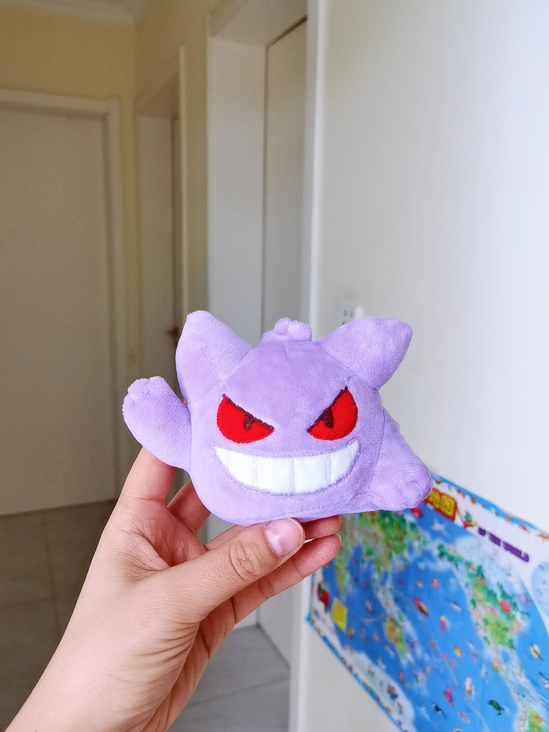 A hand holding a small purple Gengar plush toy with red eyes and a wide white grin, photographed indoors against a white wall and doorway background.