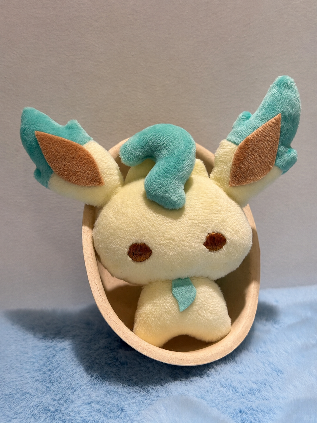 A plush toy of a Leafeon pokemon with turquoise and brown accents, sitting inside a small wooden bowl on a blue fuzzy surface against a plain gray wall.