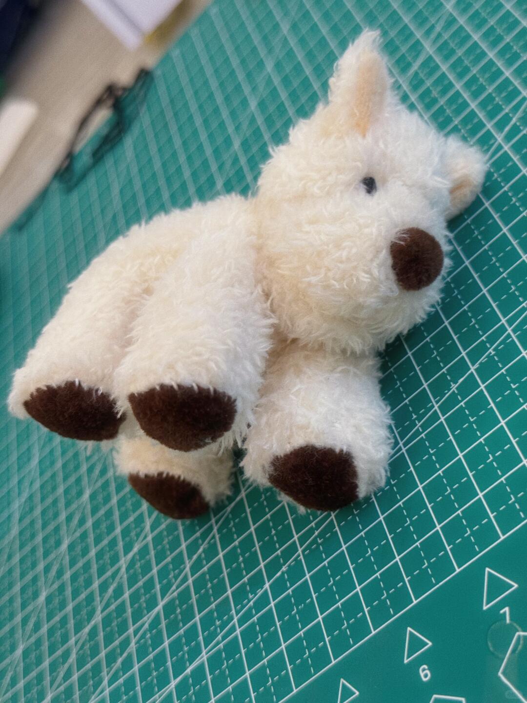 Small white plush dog toy with brown paws on a green cutting mat.
