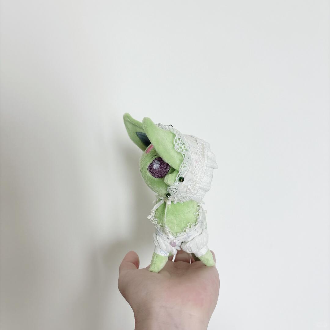 A small green pokemon eevee plush toy with a white lace bonnet and white dress, held in a person's hand against a plain light background.