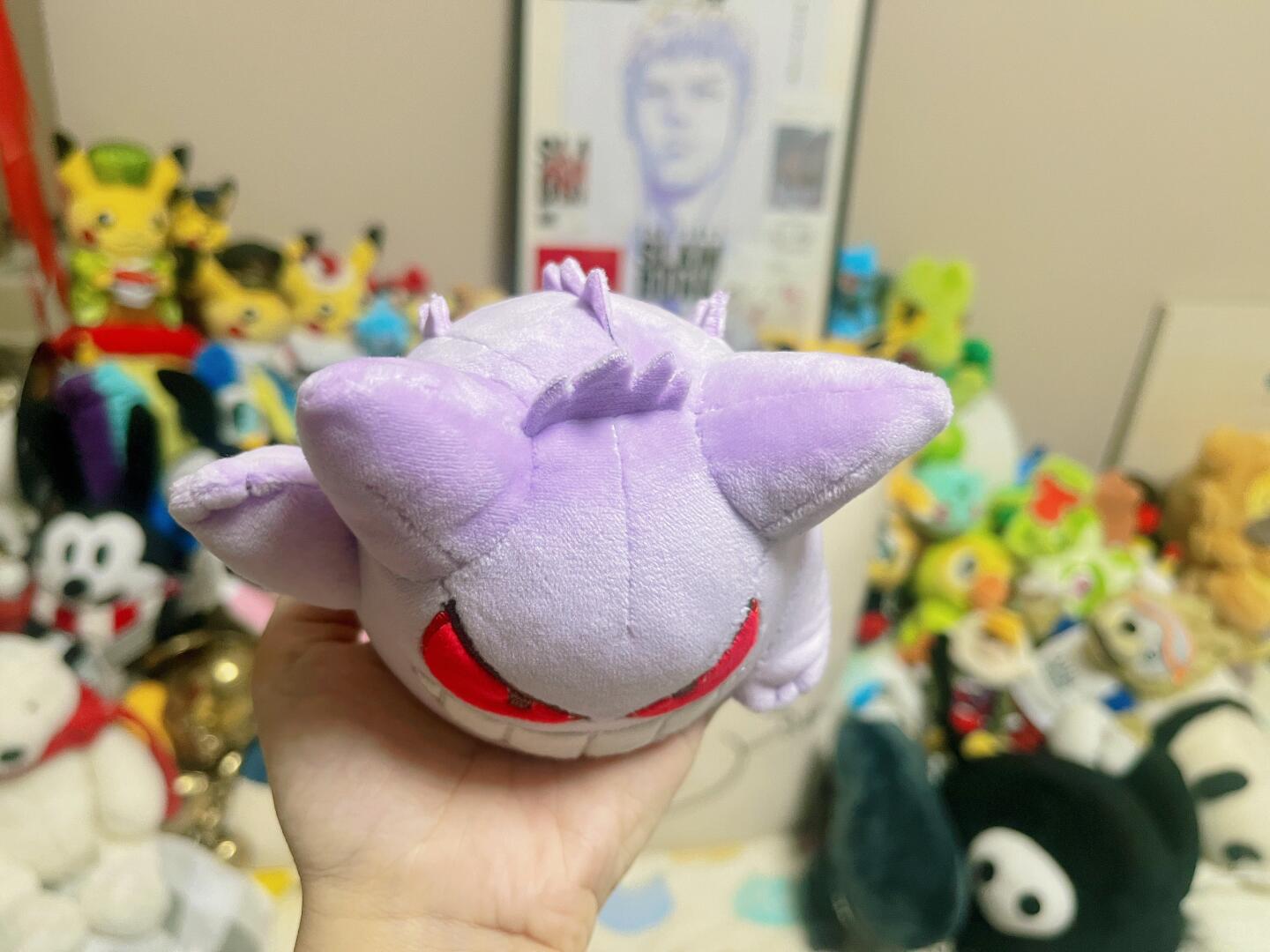A hand holding a small purple Gengar plush toy with large ears and red curved mouth details, with a collection of other plush toys in the background.