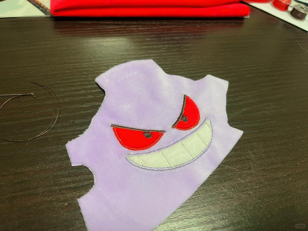 A light purple ghost-shaped Gengar fabric patch with red eyes and a white stitched grin, placed on a dark wooden tabletop.