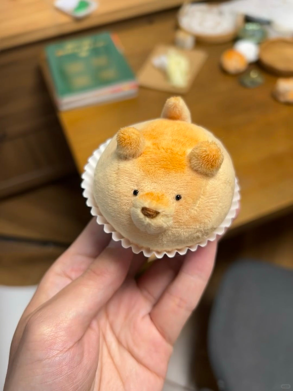 A hand holding a small plush toy shaped like a teddy bear wearing a cupcake, with a wooden table and blurred objects in the background.