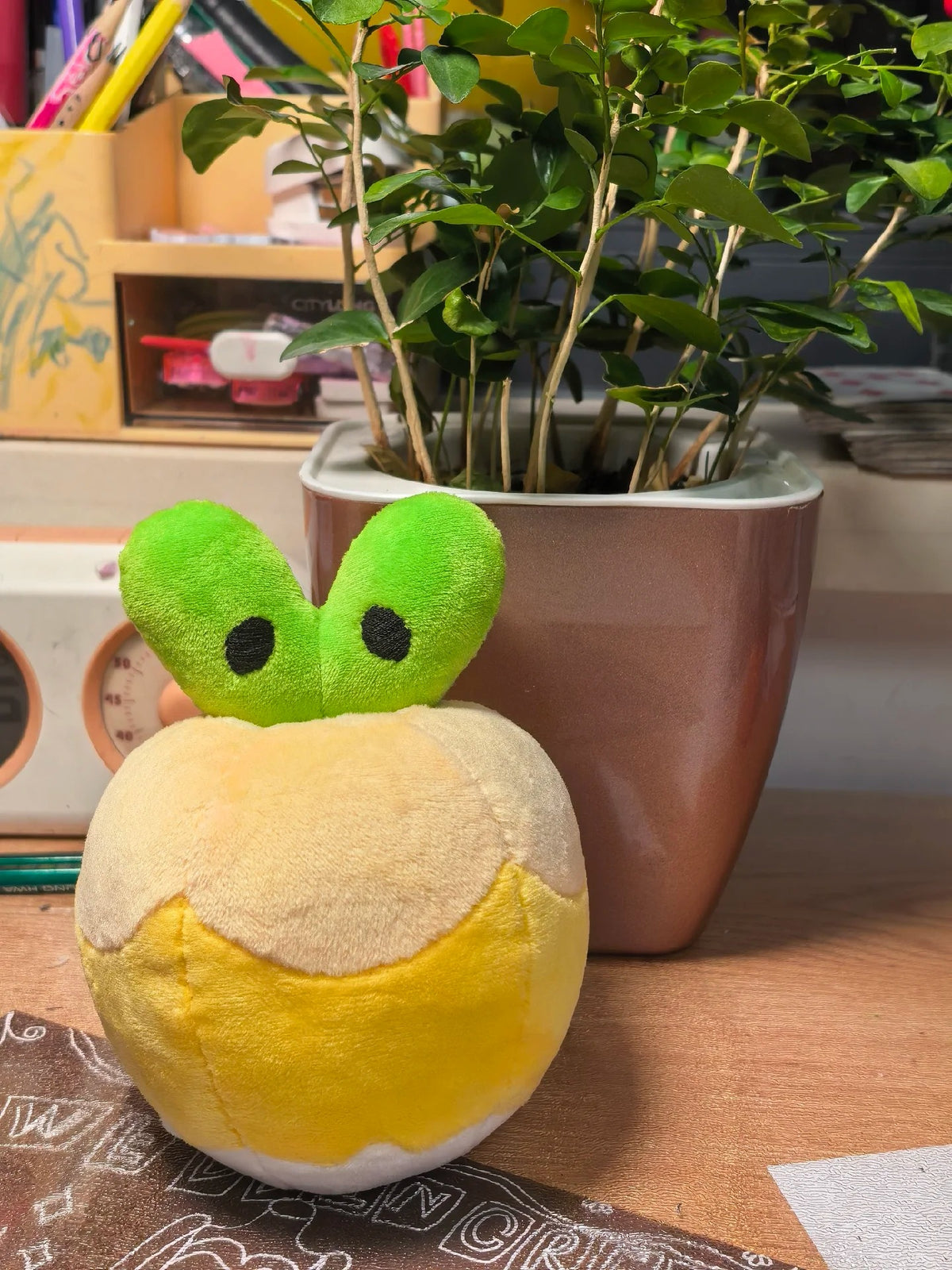A Applin plush toy shaped like a cracked yellow-and-beige apple with a green leafy top and black eyes, placed on a wooden surface in front of a potted plant.