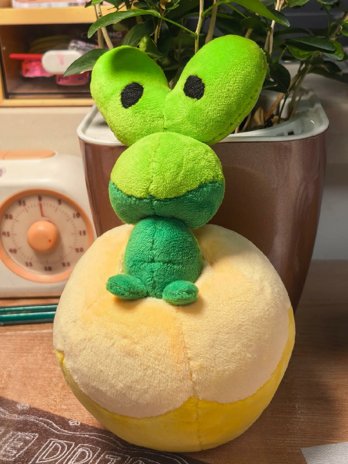 A Applin plush toy featuring a bright green frog character sitting on a round yellow and cream-colored base, placed on a wooden surface in front of a potted plant.