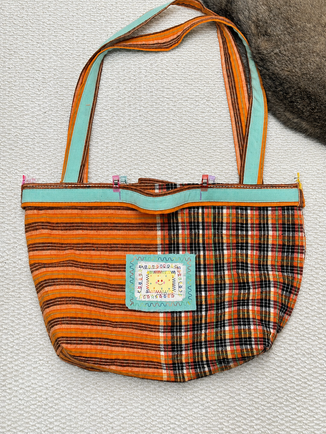 A handmade fabric shoulder bag with a curved bottom, featuring orange, black, and beige striped fabric on one side and a black, orange, and beige plaid pattern on the other. It has a light blue-green trim and matching shoulder straps, with a decorative embroidered patch on the front.