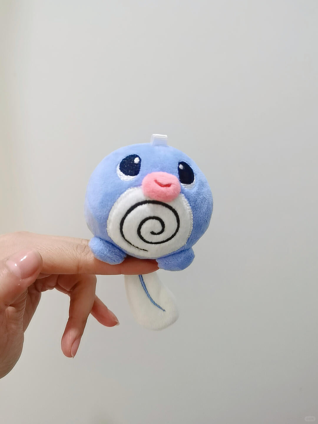 A small blue Poliwag plush character keychain or bag charm with a pink nose and a white spiral belly, held by a person's hand against a plain light gray background.