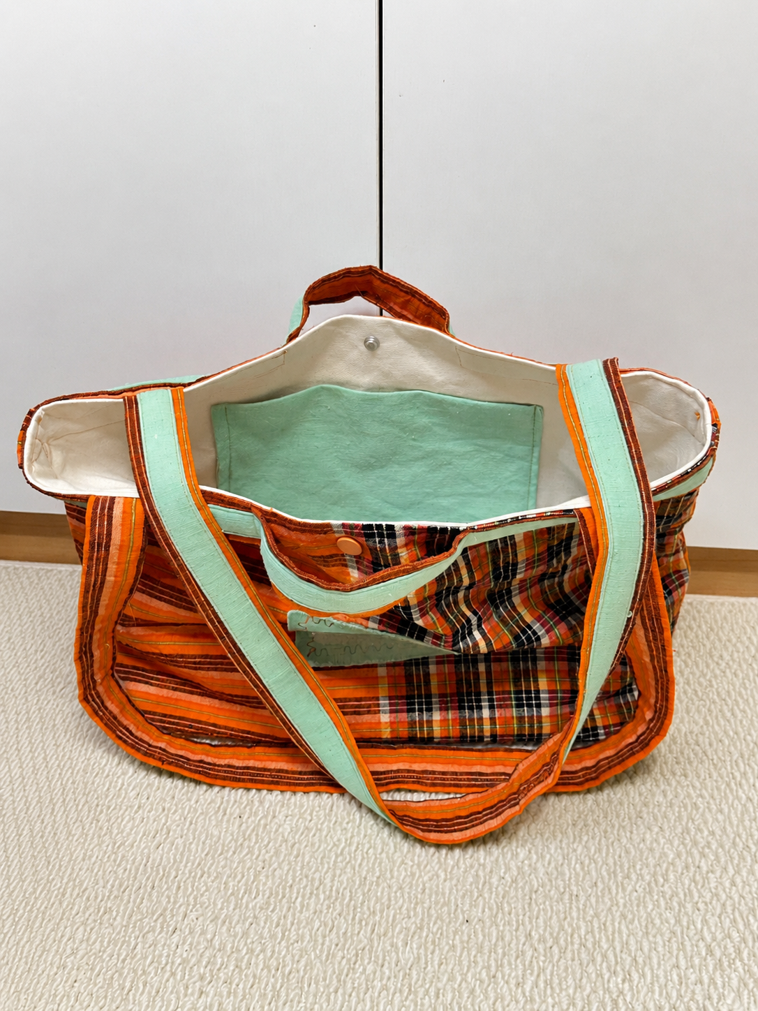 A handmade tote bag made from orange and black plaid fabric with mint green trim and handles, shown on a light beige carpet against a white wall.
