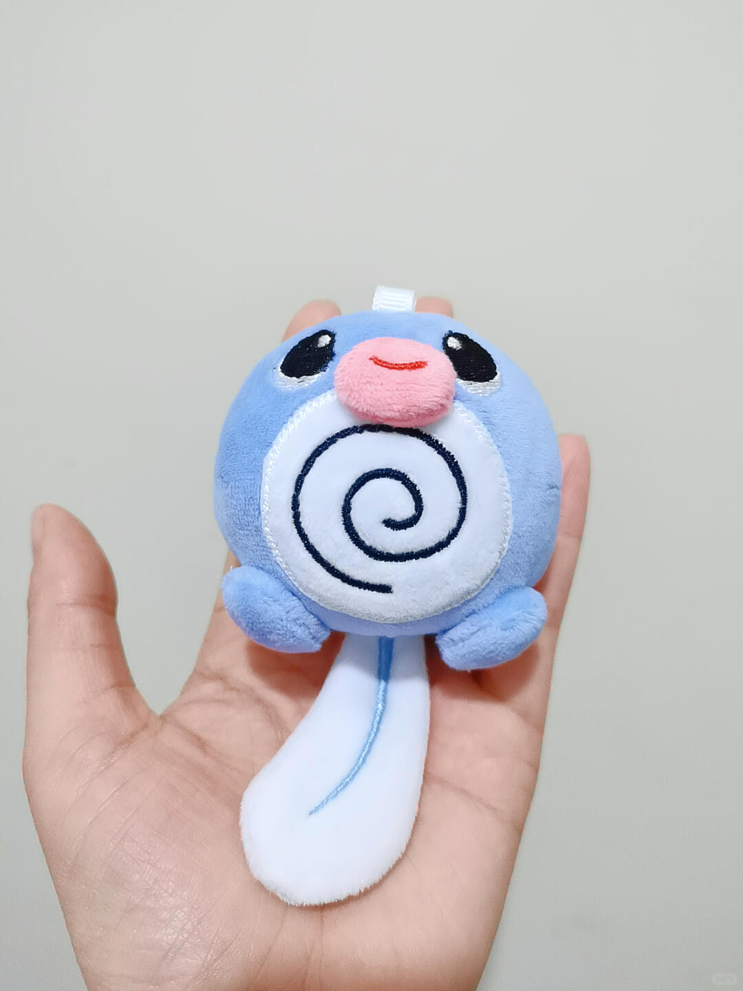 A small plush toy Poliwag character with a light blue body, white underside, black spiral face detail, and a pink nose, held in a person's hand against a plain light background.
