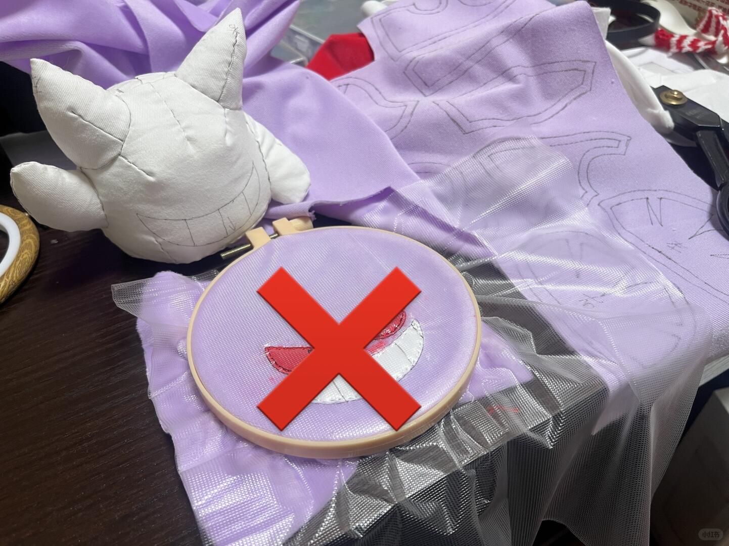 A purple embroidered Gengar fabric hoop with a partially stitched character design, placed on a table with lavender fabric, a white plush toy, and a large red X overlay covering the center.