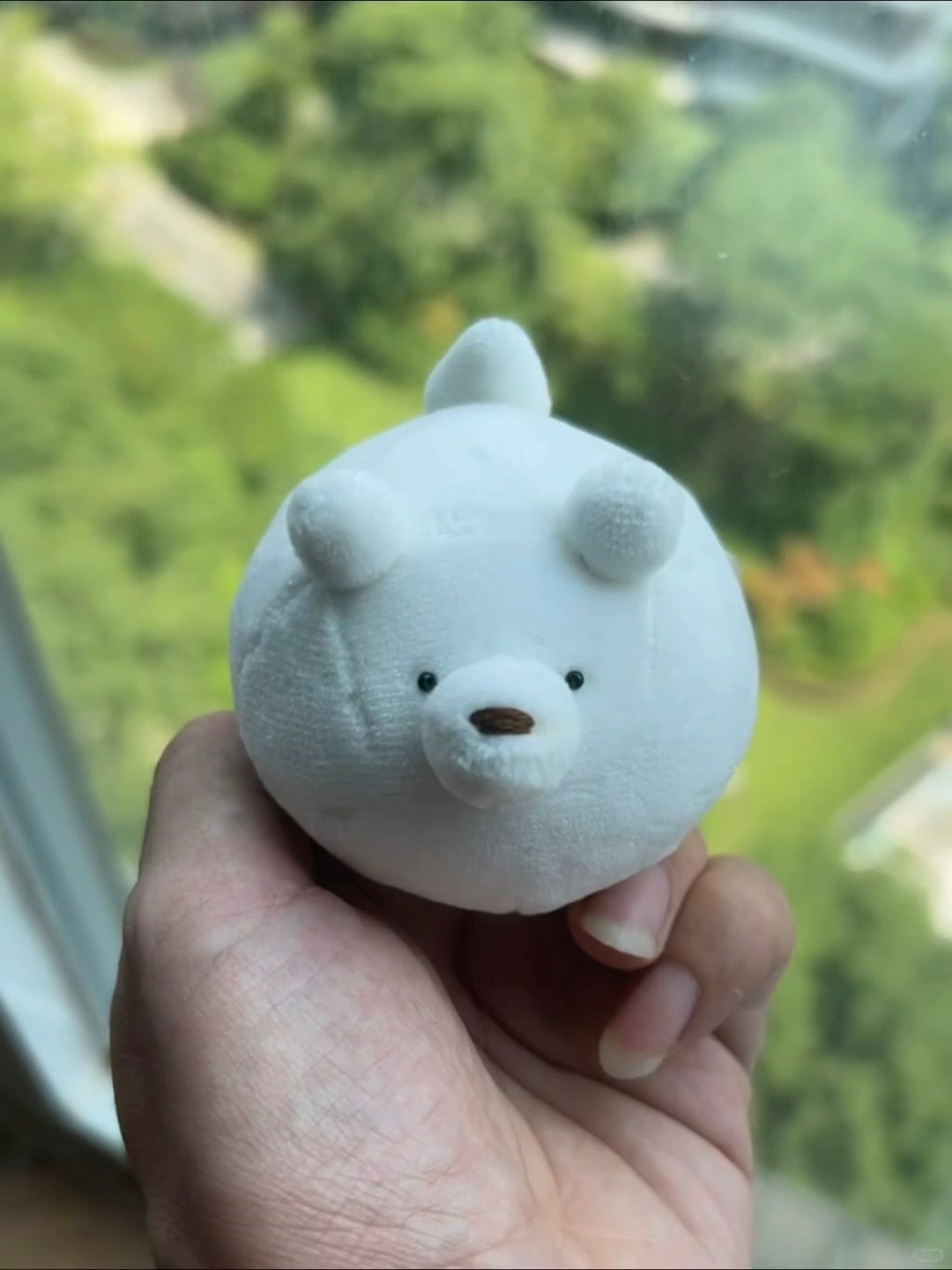 A small white plush bear toy with embroidered black eyes and nose, held in a person's hand in front of a blurred window view of green trees.