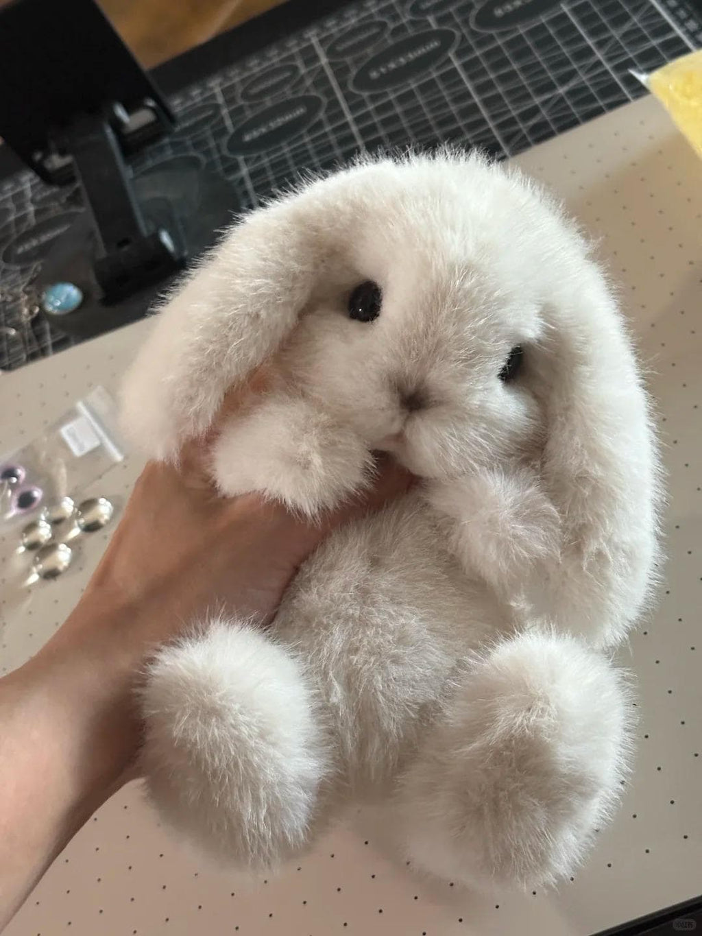 White fluffy toy held by a hand on a cutting mat with buttons nearby