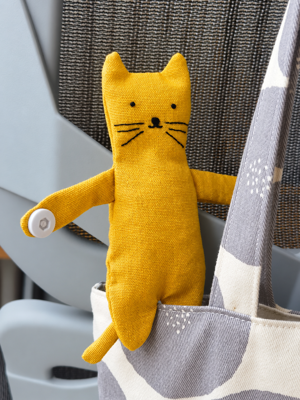 A yellow fabric cat-shaped plush toy with embroidered facial features and a white button on one paw, hanging from the strap of a gray and white patterned tote bag.