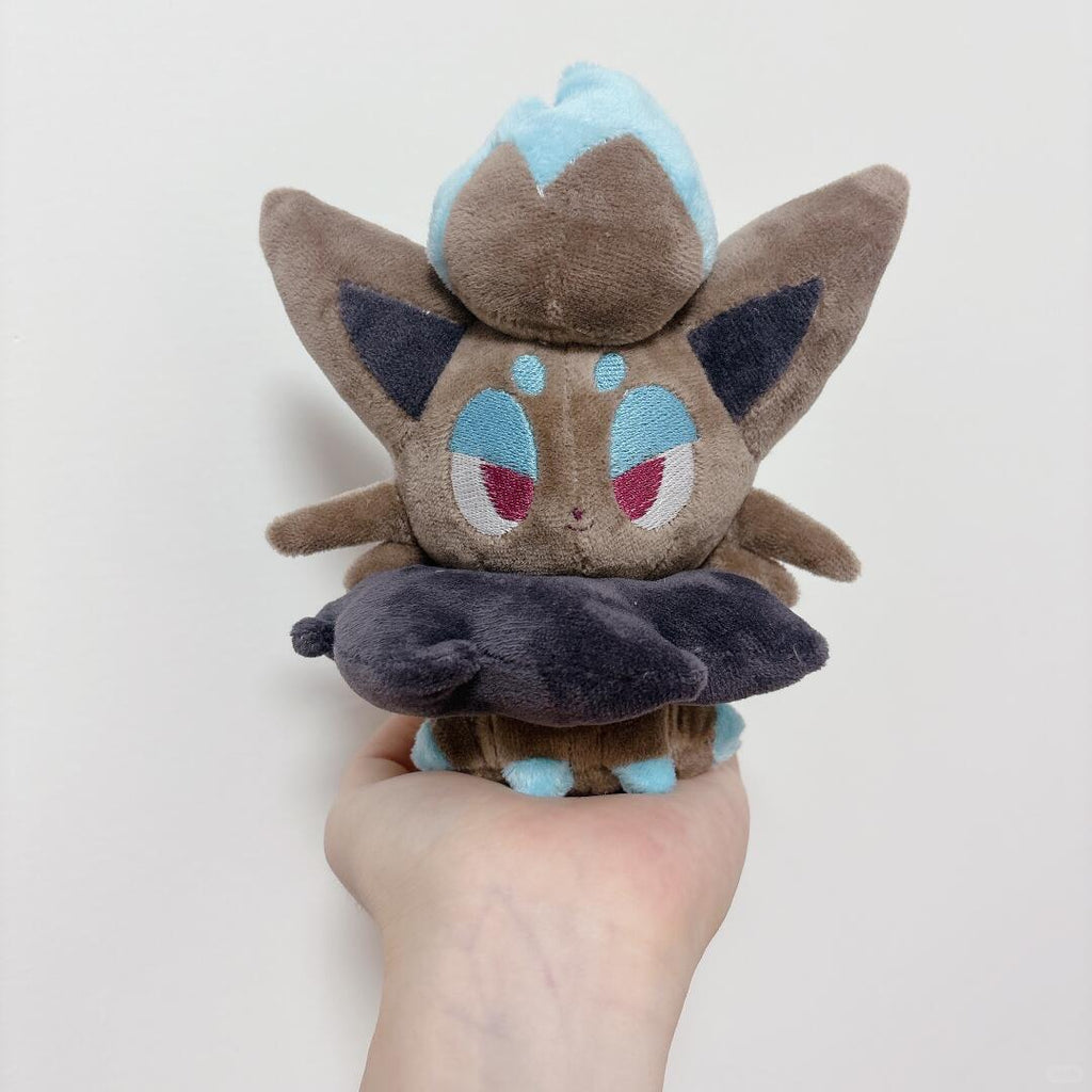 A small plush zorua toy held in a person's hand against a plain light background. The toy is brown with light blue and dark blue accents, resembling a cute character plush.