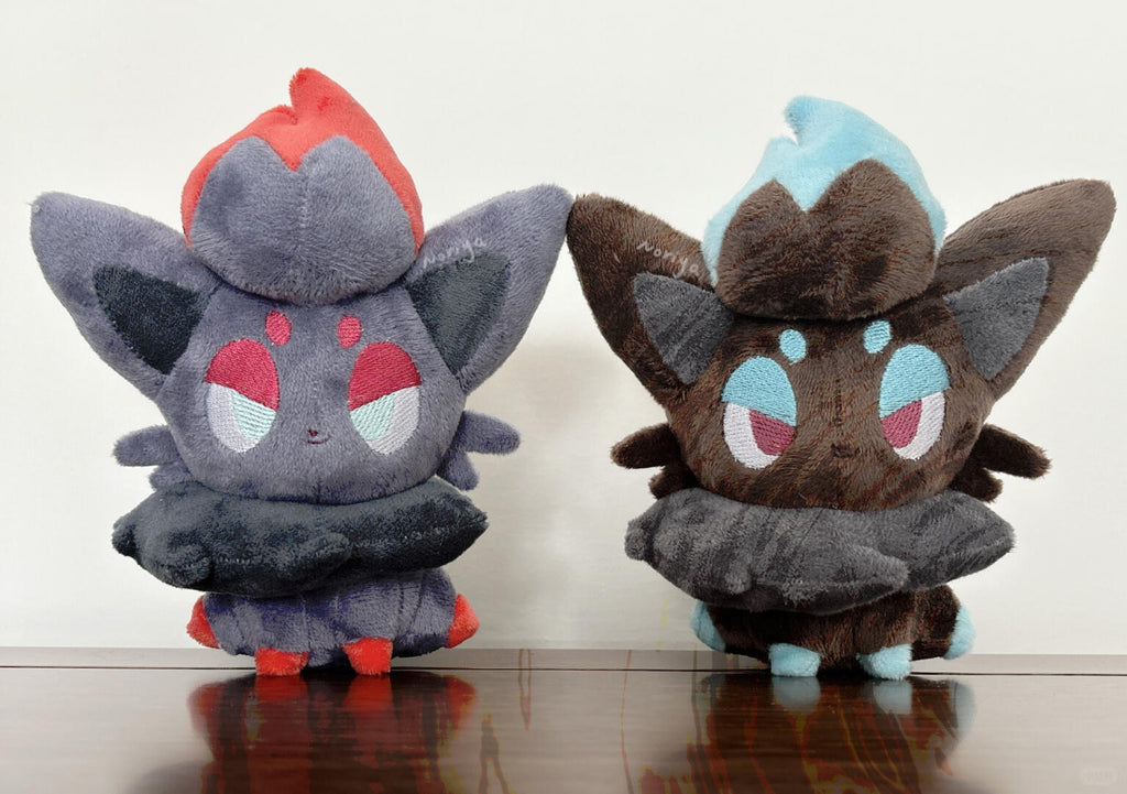 Two small plush toy zorua characters sitting side by side on a dark wooden surface against a plain light background. The left plush is dark gray with red and light blue accents, and the right plush is dark brown with light blue and pink accents.