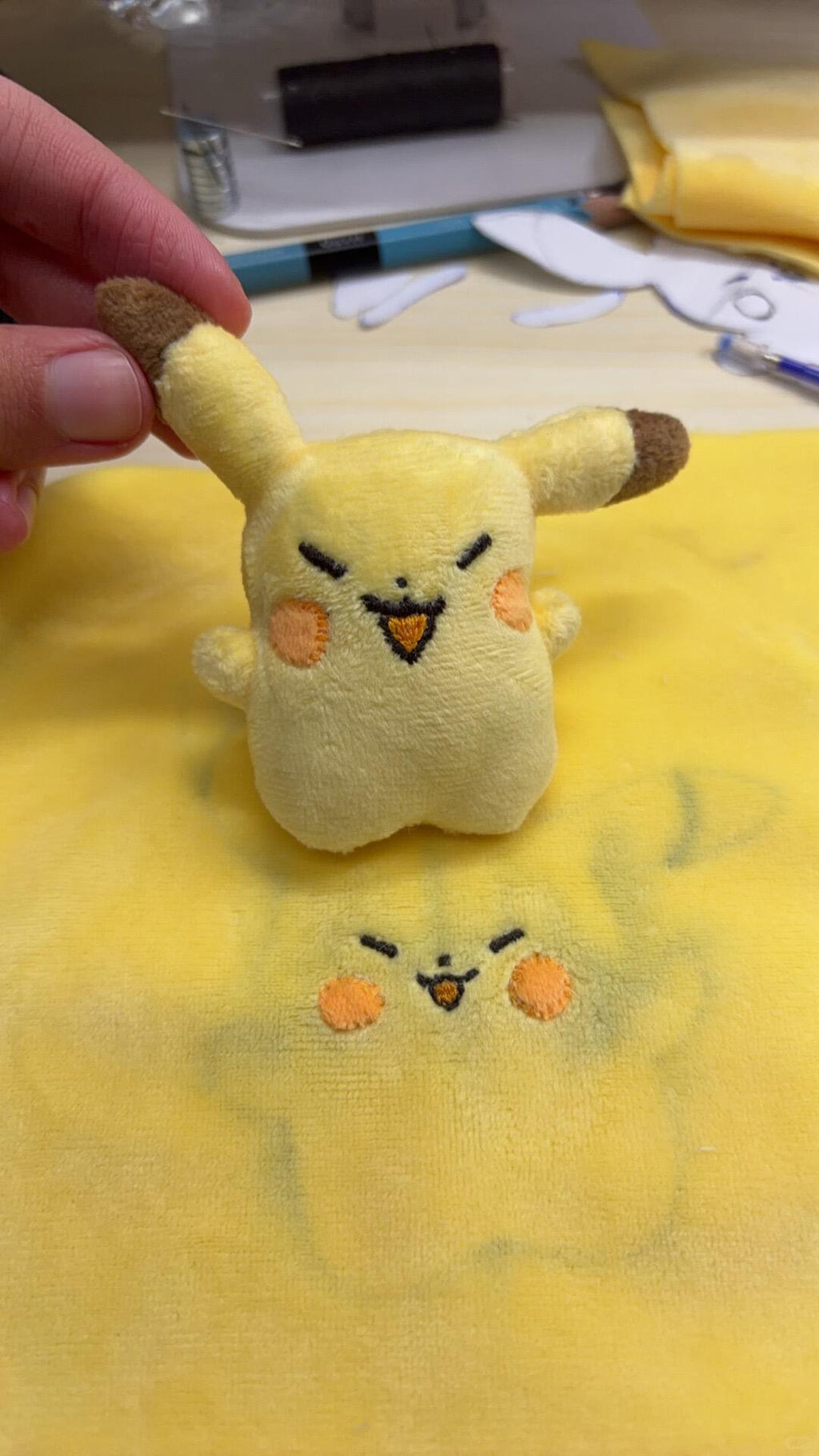 Small plush toy resembling Pikachu held over a larger yellow fabric with similar design.