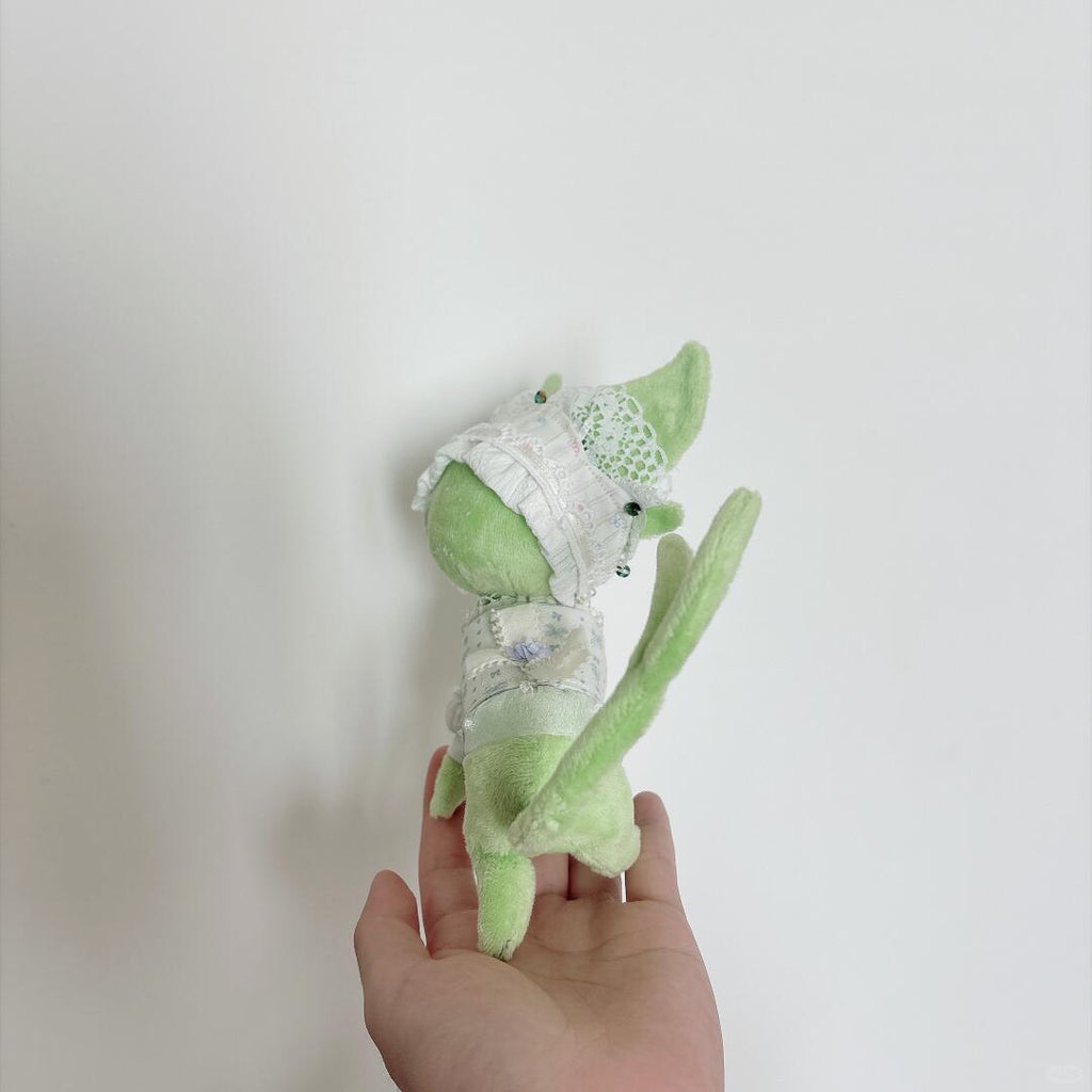 A small light green pokemon eevee plush toy with a long tail and white lace-like clothing, held in a person's hand against a plain light background.