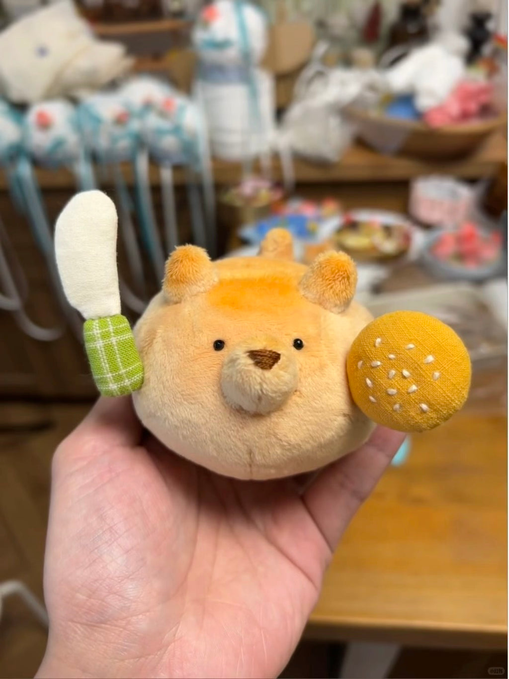 A small plush toy held in a person's hand, featuring a round tan body with embroidered facial features, a white and green plaid tail, and a yellow bun-shaped accessory with white dots.