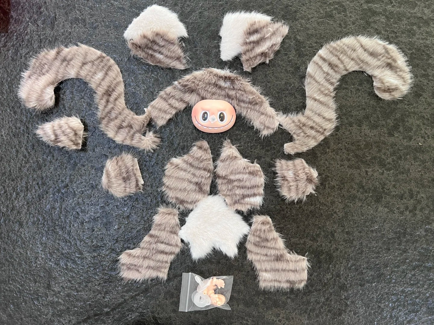 Labubu cat sewing pattern. Fur-like material pieces on a dark surface with a small pink object in the center. 