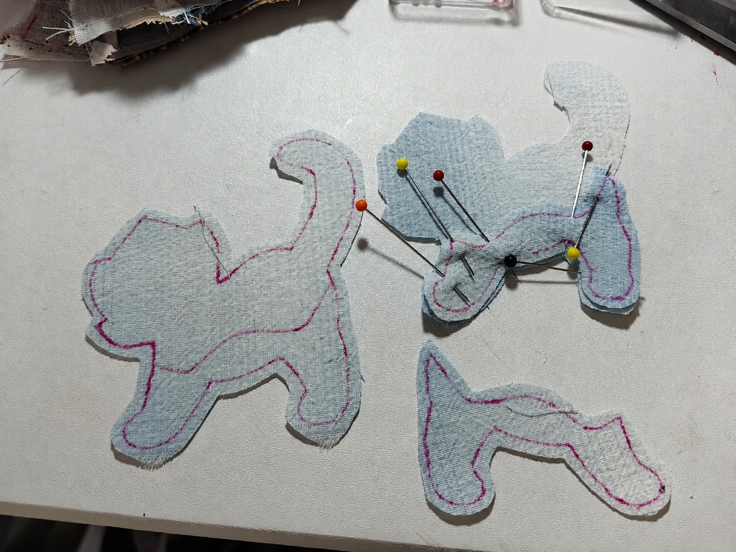 Three light blue fabric dog-shaped pieces with pink stitched outlines, pinned together on a white surface.