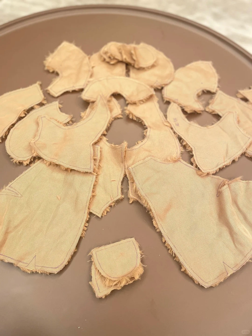 A set of Jellycat teddy bear sewing pattern, beige fabric garment pieces, likely doll or costume bodysuit sections, laid out on a brown plate.