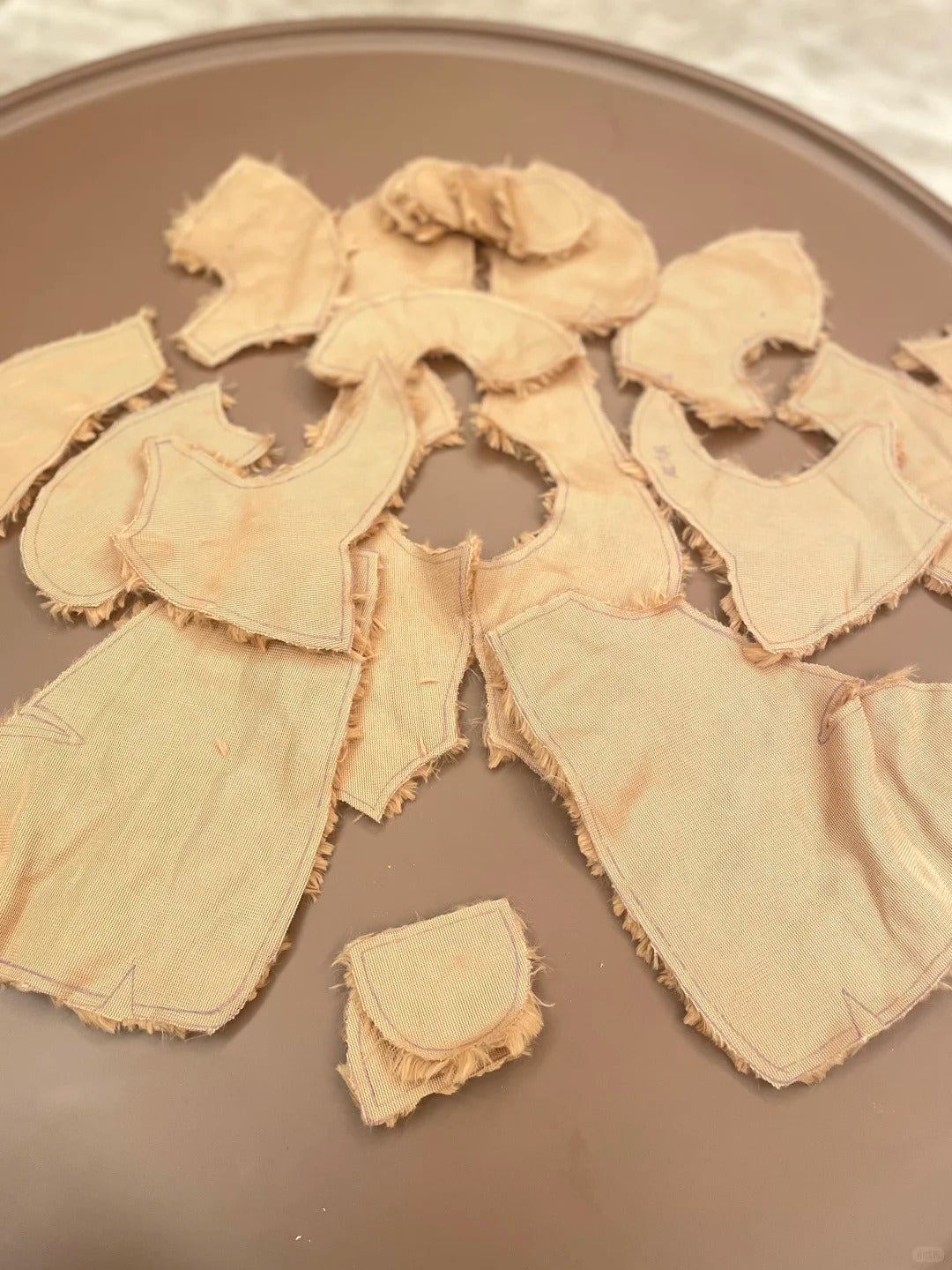 A set of Jellycat teddy bear sewing pattern, beige fabric garment pieces, likely doll or costume bodysuit sections, laid out on a brown plate.
