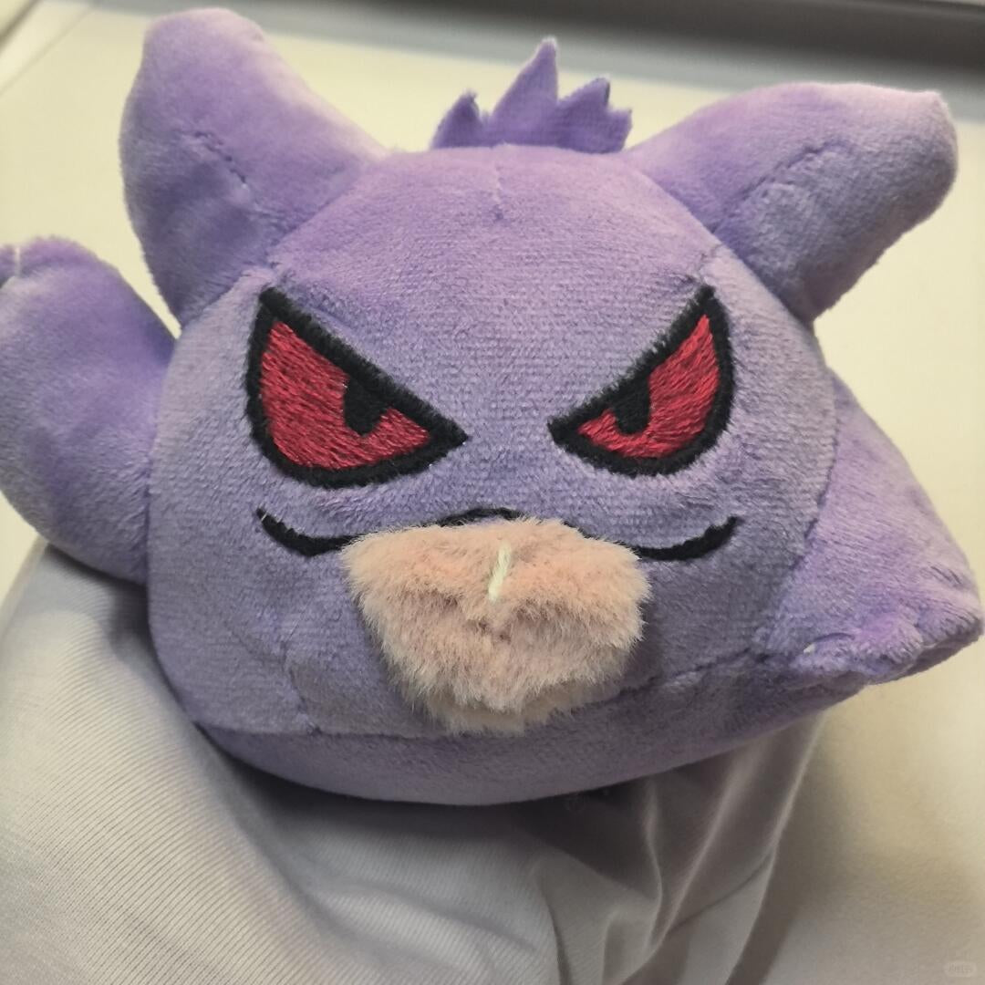 A purple Gengar plush toy with red embroidered eyes and a beige fuzzy snout, photographed close up against a light neutral background.