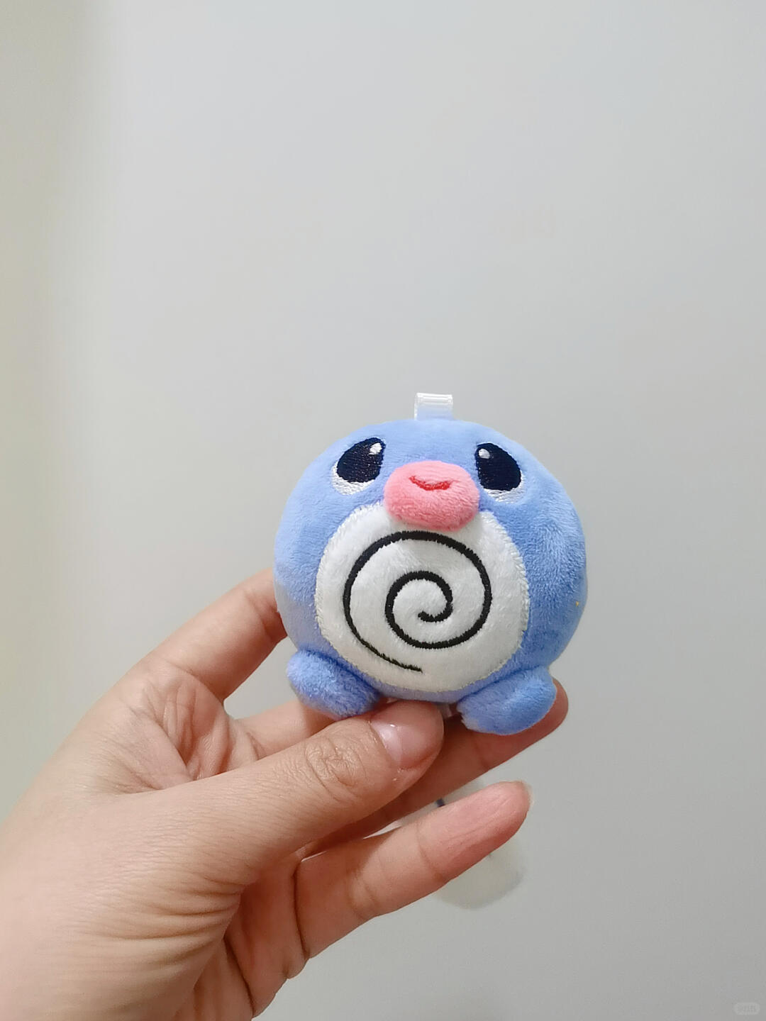 A small Poliwag plush toy character held in a person's hand against a plain light gray background. The character is light blue with a white spiral chest design, black eyes, and a pink nose.