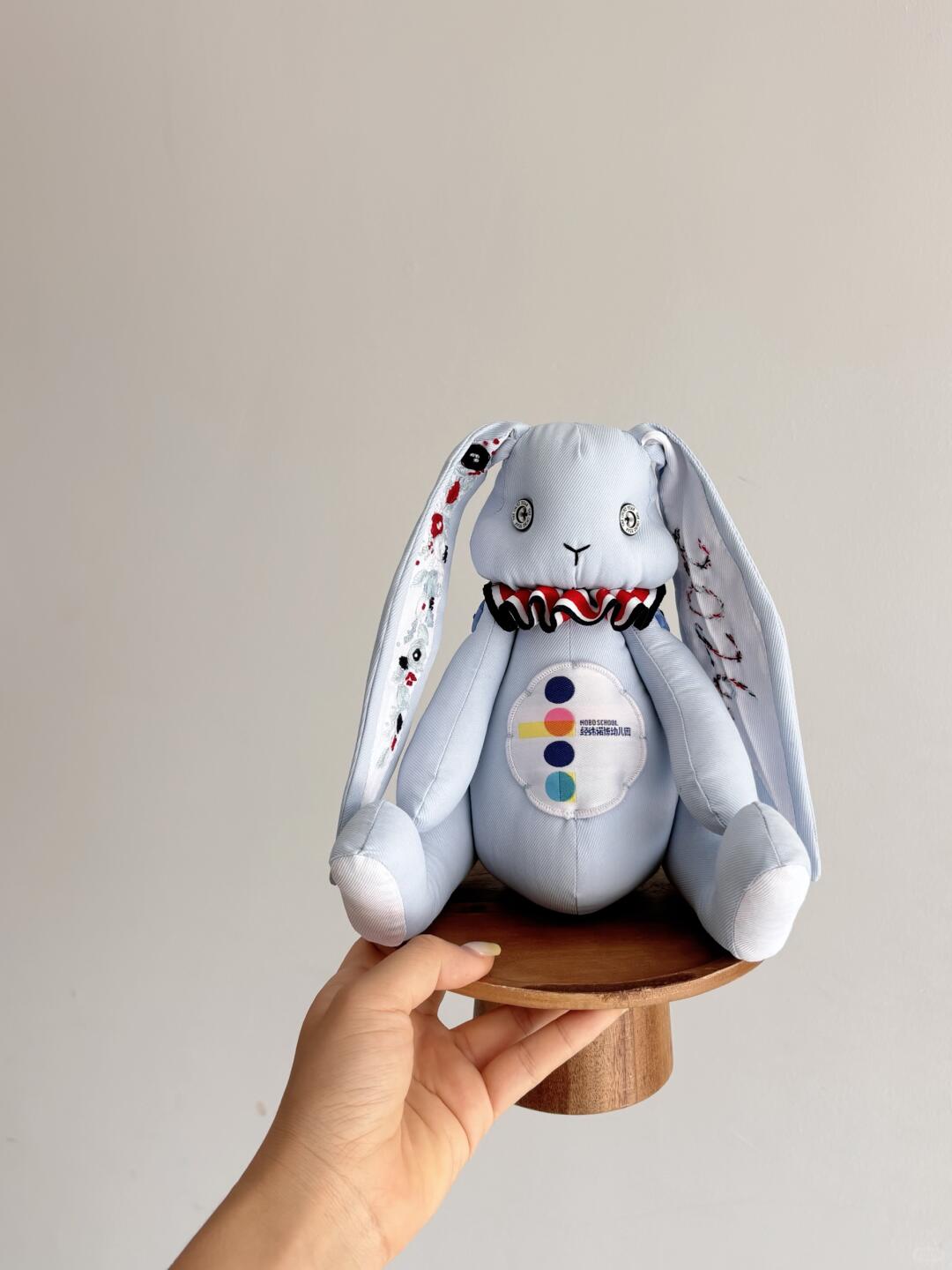Light blue denim bunny plush seated on a small wooden stand, with embroidered ears and a ruffled collar.