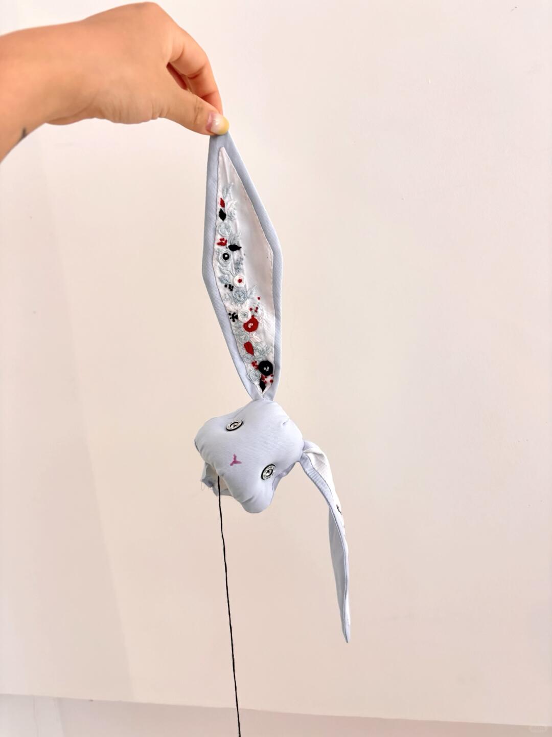 Light blue denim bunny plush suspended by one embroidered ear, showing its long limbs and stitched face.