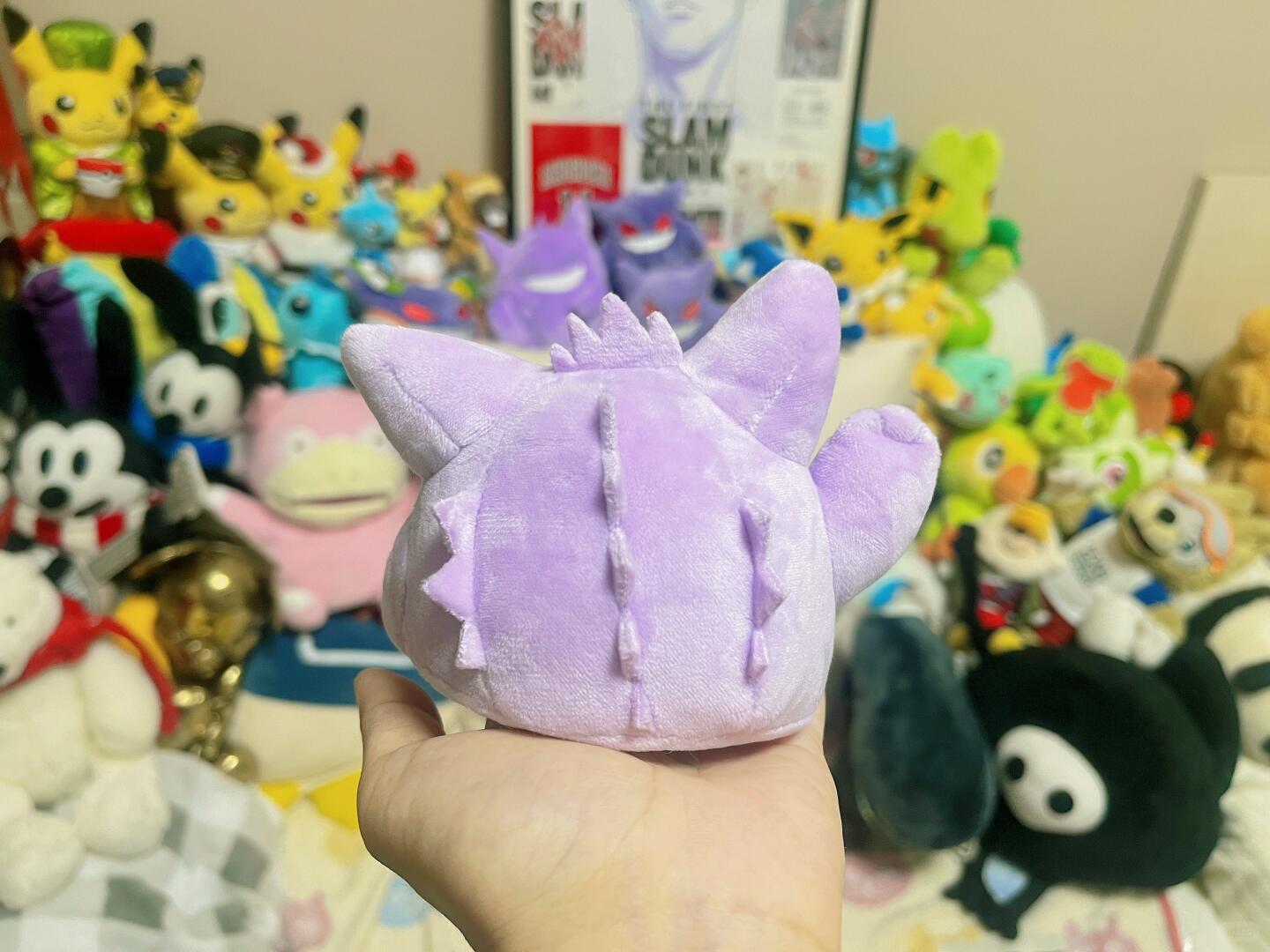 A hand holding a small purple Gengar plush toy with pointed ears and spiky details, set against a background filled with various colorful plush toys and collectibles.