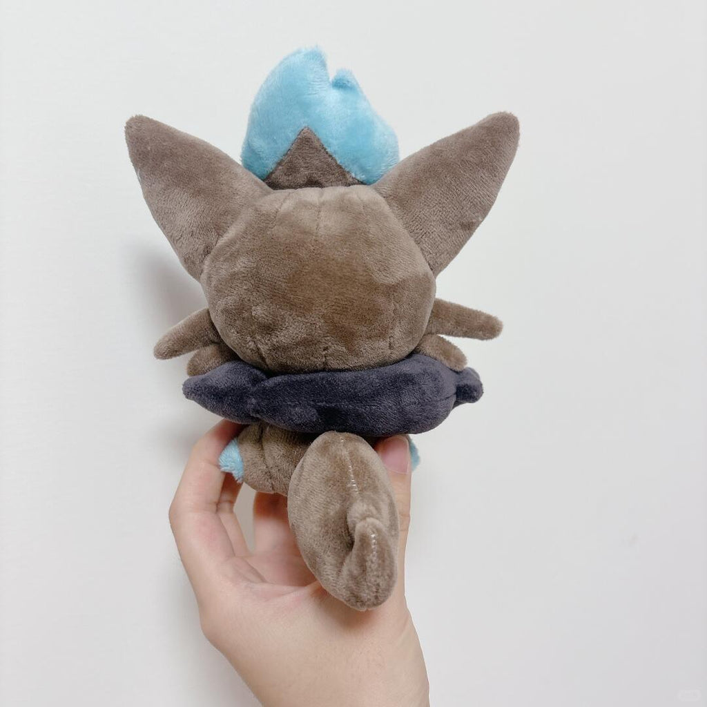 A zorua plush toy held in a person's hand against a plain light background. The toy appears to be a small brown animal character with large pointed ears, a light blue tuft on top, and a dark navy ring around the middle.